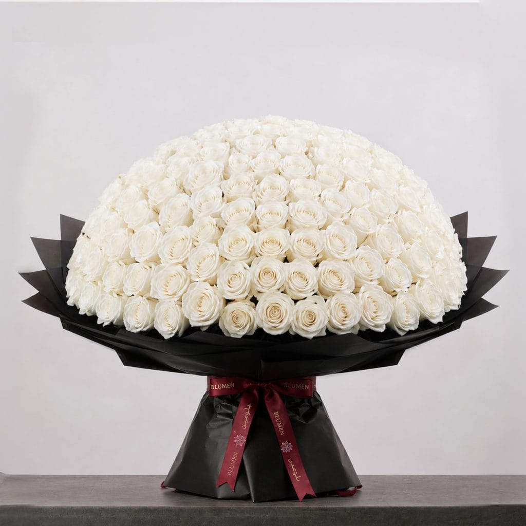 A large bouquet of elegant white roses arranged in a full, rounded shape, wrapped in sophisticated black paper and finished with a refined ribbon.