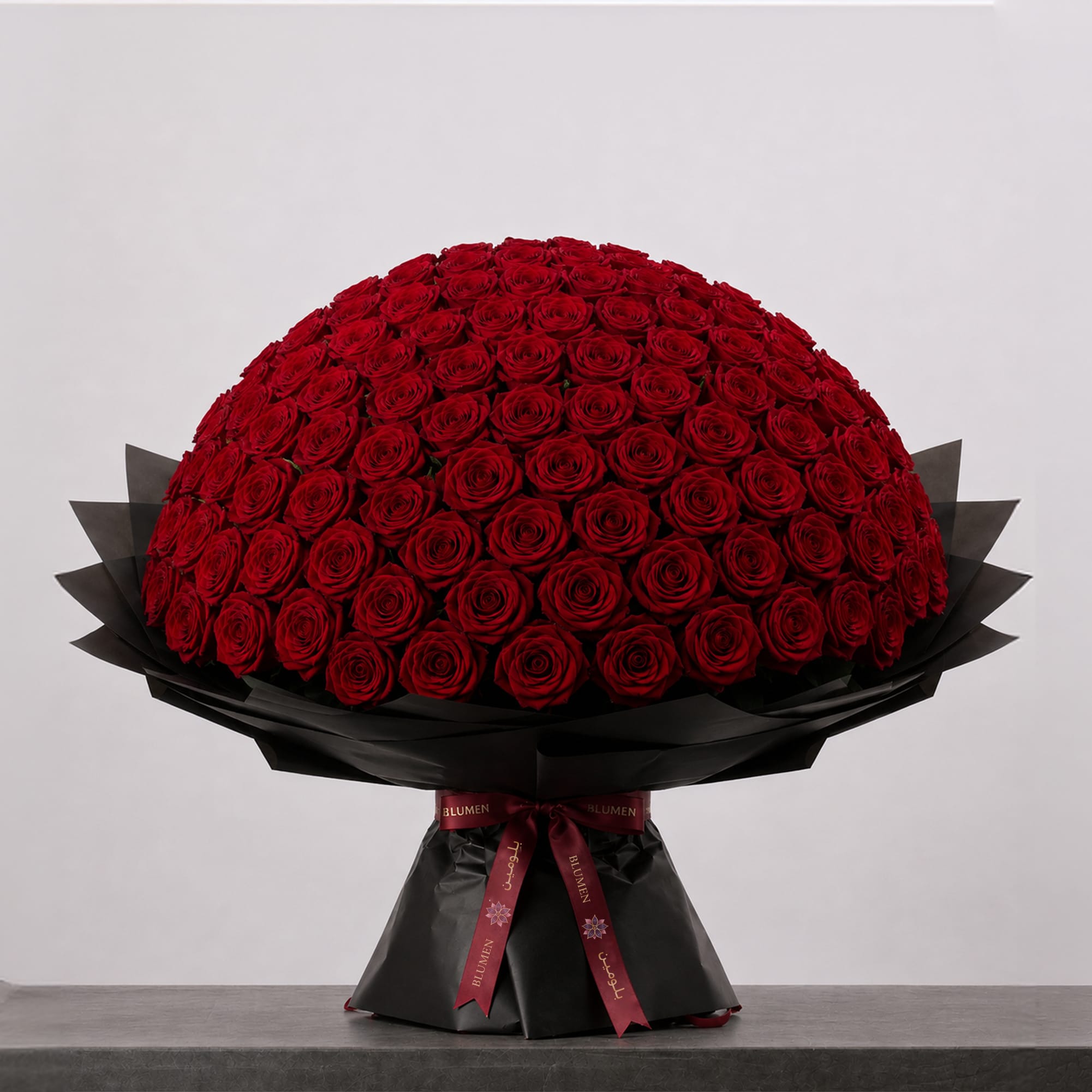 A large bouquet of deep red roses arranged in a full, rounded shape, wrapped in elegant black paper and finished with a burgundy ribbon.