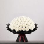 A large bouquet of elegant white roses arranged in a full, rounded shape, wrapped in sophisticated black paper and finished with a refined ribbon.