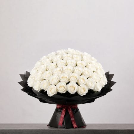 A large bouquet of elegant white roses arranged in a full, rounded shape, wrapped in sophisticated black paper and finished with a refined ribbon.