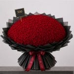 A large bouquet of deep red roses arranged in a full, rounded shape, wrapped in elegant black paper and finished with a burgundy ribbon.