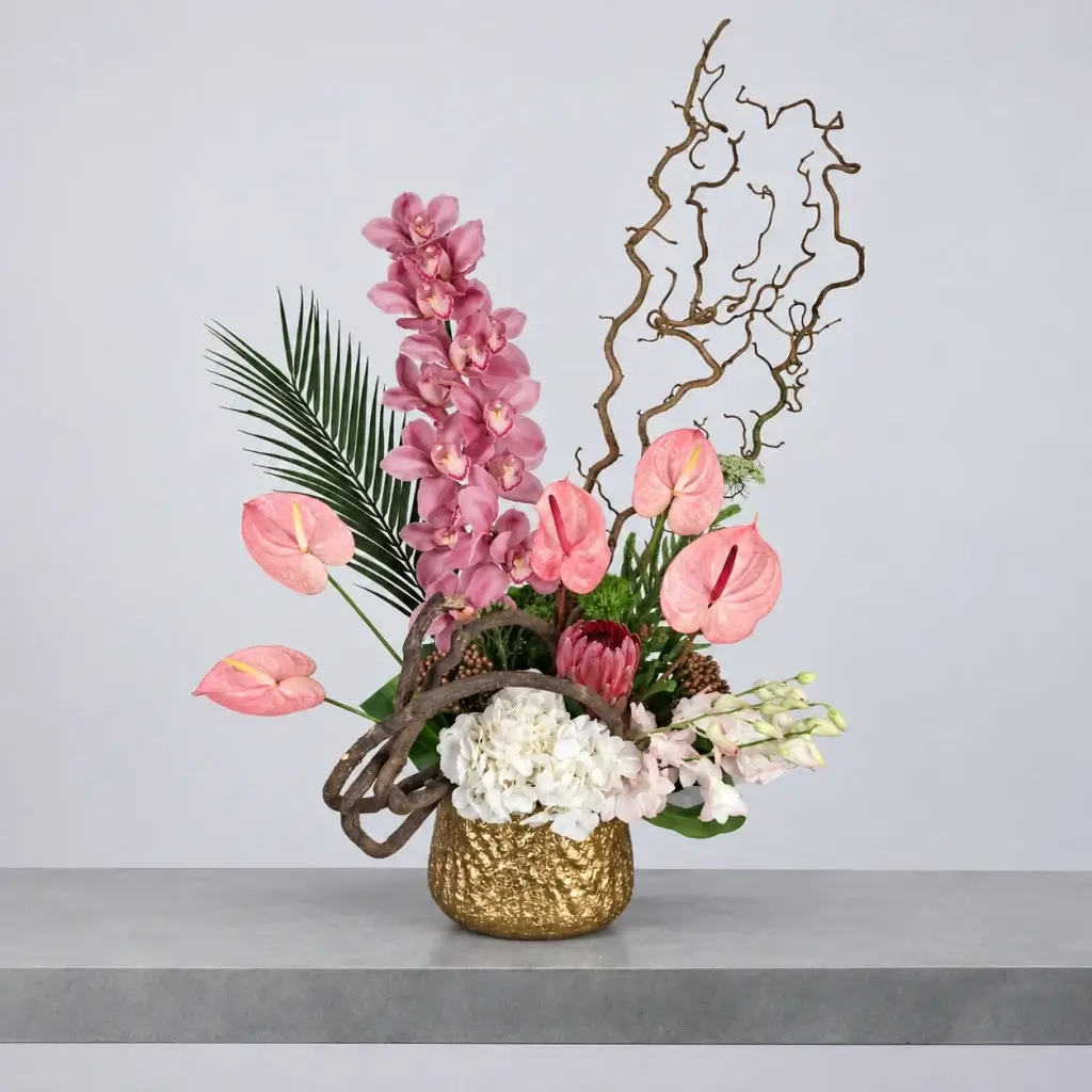 Rosé d’Or luxury floral arrangement featuring pink Cymbidium orchids, pink anthuriums, white hydrangea, protea and sculptural branches in an elegant textured gold vase by Blumen.